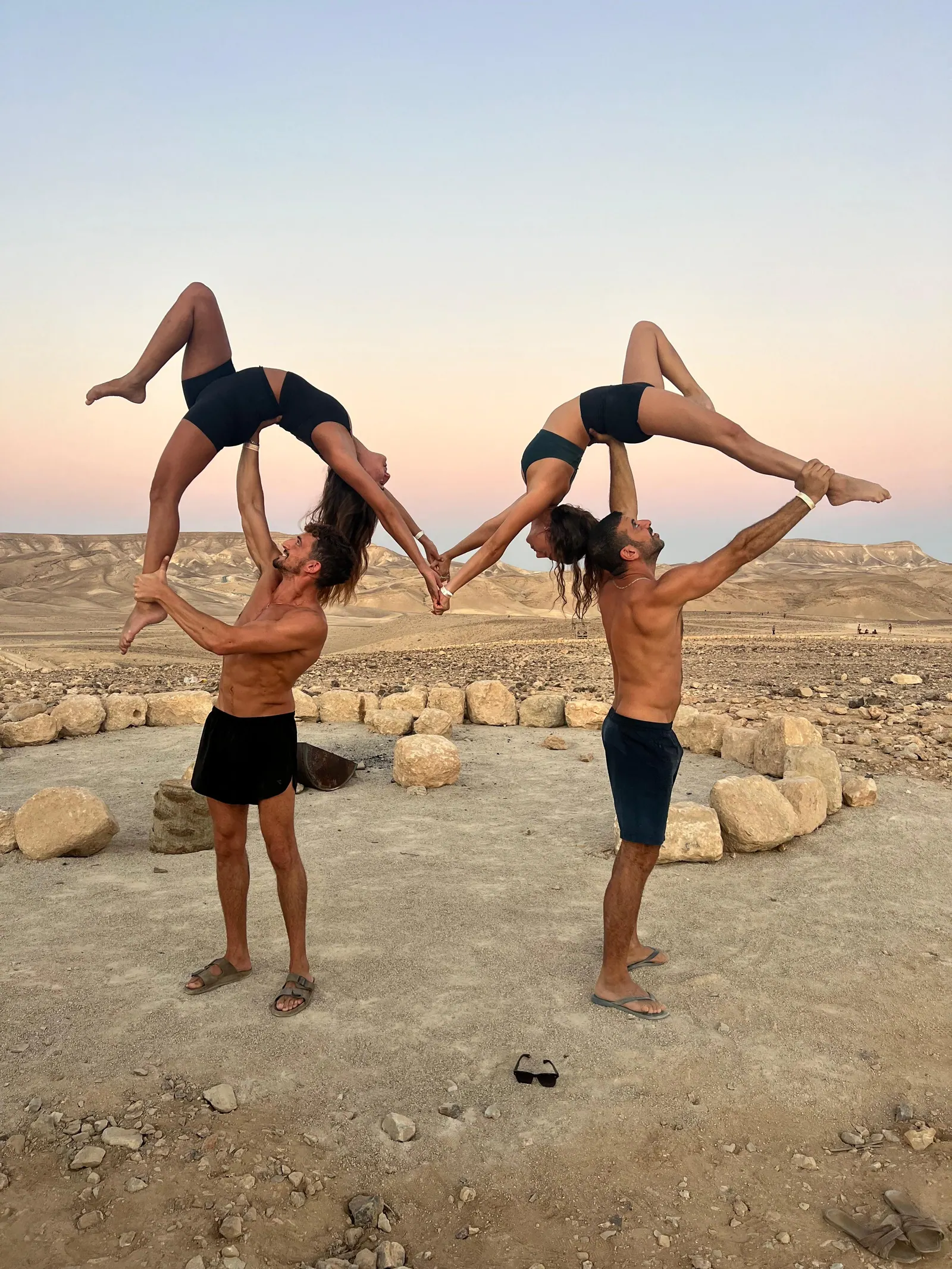 שני זוגות אקרויוגה בתנוחה מסונכרנת במדבר הנגב בשקיעה — ארבעה מתרגלים, פלייריות אוחזות ידיים באוויר